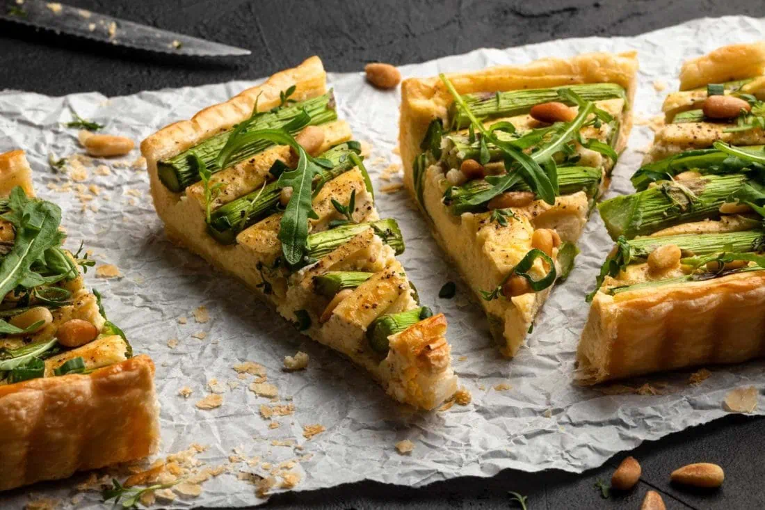 Grüne Spargeltarte mit Blätterteig in Stücke geschnitten auf einem weißen Papier. Auf der Tarte liegen Pinienkerne und Rucola.