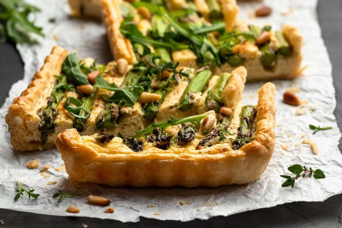 Spargeltarte mit grünem Spargel in Stücke geschnitten auf einem Papier. Garniert mit Kräutern, Pinienkernen und Rucola.