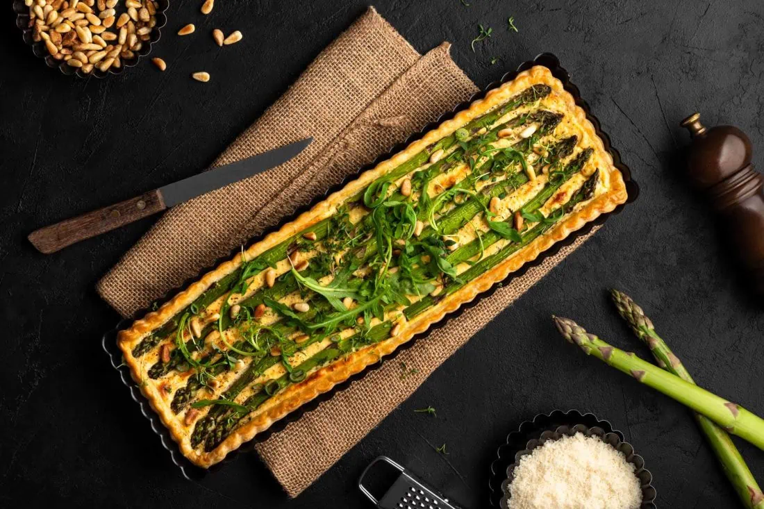 Feine grüne Spargeltarte mit Blätterteig in einer länglichen Tarteform. Daneben stehen Pinienkernen und Parmesan.