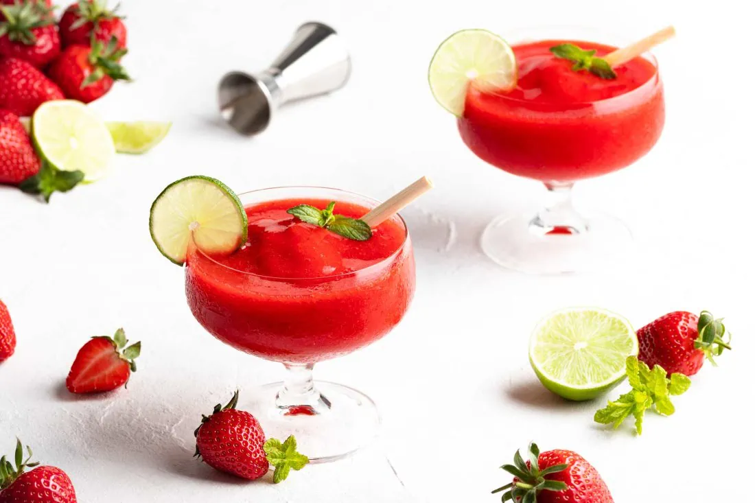 Frozen Erdbeer Daiquiris in Gläsern mit Stiel an denen sich Kondenswasser bildet. Daneben frische Erdbeeren und Limette.