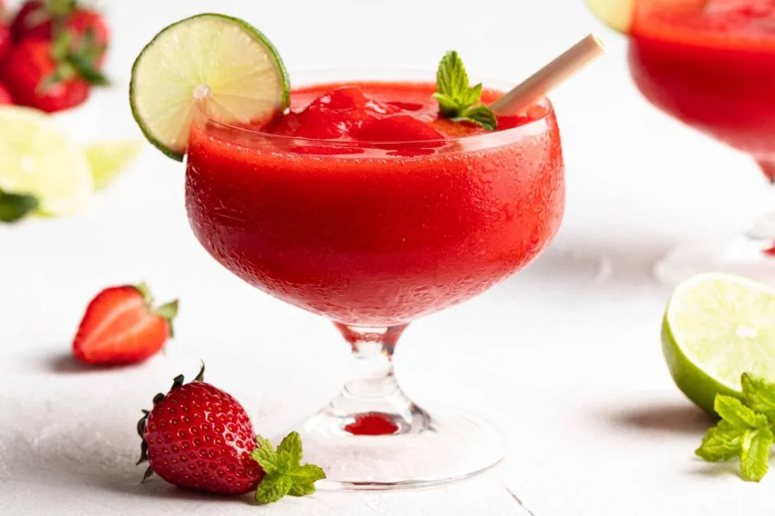 Frozen Erdbeer Daiquiri in einem eiskalten Glas. Daneben liegen frische Erdbeeren, Minze und Limetten.