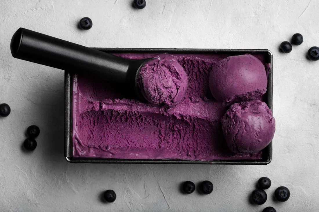 Eine rechteckige Form gefüllt mit fruchtigem Blaubeereis in der Farbe Lila. Auf dem Eis liegen mehrere Kugeln und eine Eisportionierer.