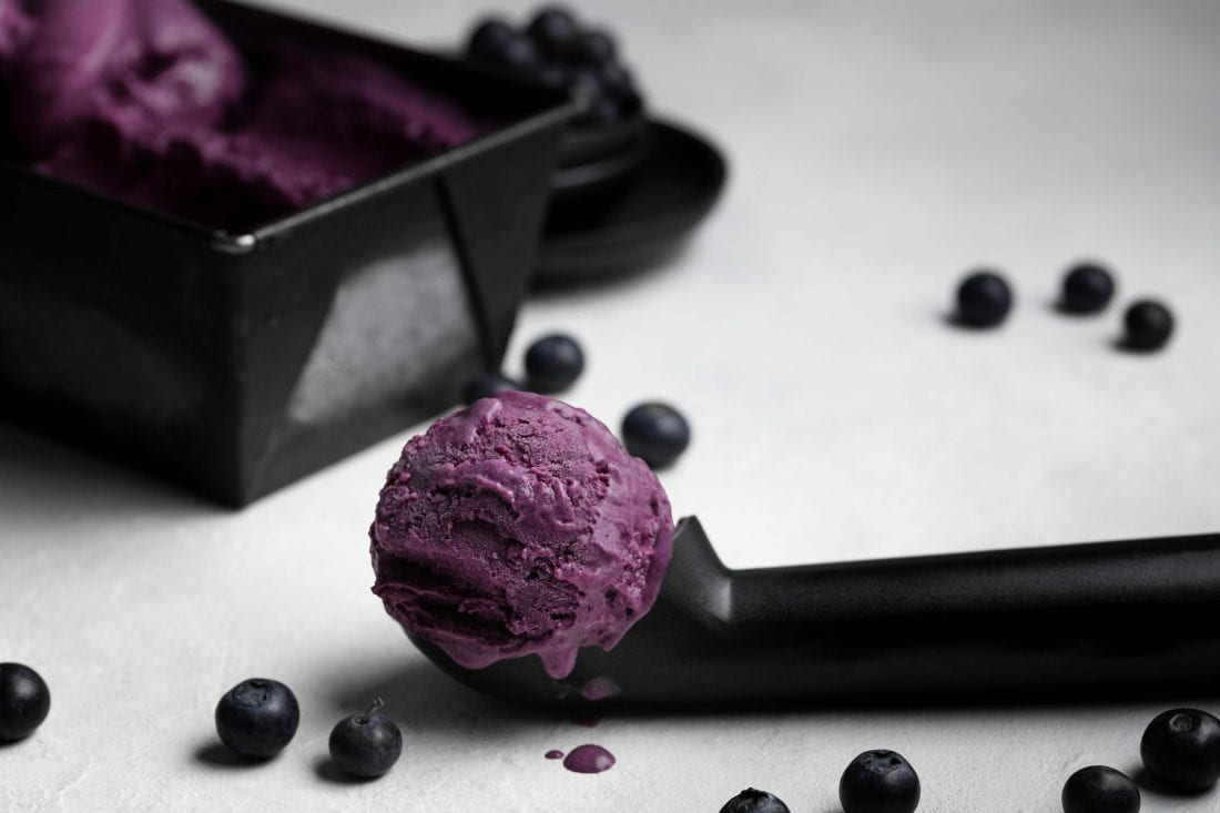 Eine Kugel lilafarbenes, fruchtiges Blaubeereis auf einem schwarzen Eislöffel.