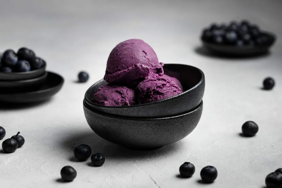 Mehrere Kugeln Blaubeereis als Eisbecher serviert in einem schwarzen Schälchen.