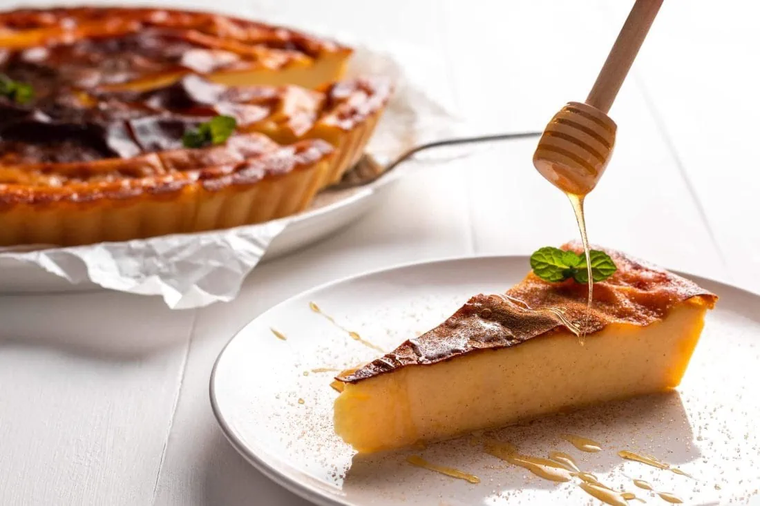 Ein Stück Galatopita auf einem Kuchenteller wird mit Honig beträufelt. Im Hintergrund steht der restliche griechische Kuchen.