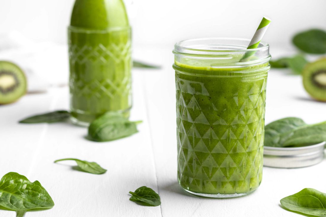 Gesunder grüner Smoothie mit Spinat, Banane und Mango in einem Glas. Hinter dem Glas steht eine Flasche gefüllt mit dem grünen Smoothie.