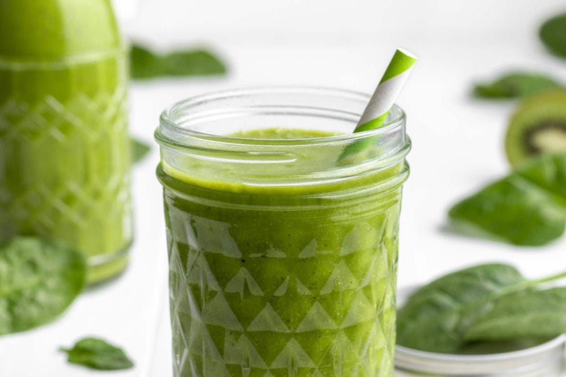 Grüner Smoothie mit Spinat in einem Schraubglas und in einer Bügelflasche. Neben dem Glas liegen mehrere frische Spinatblätter.