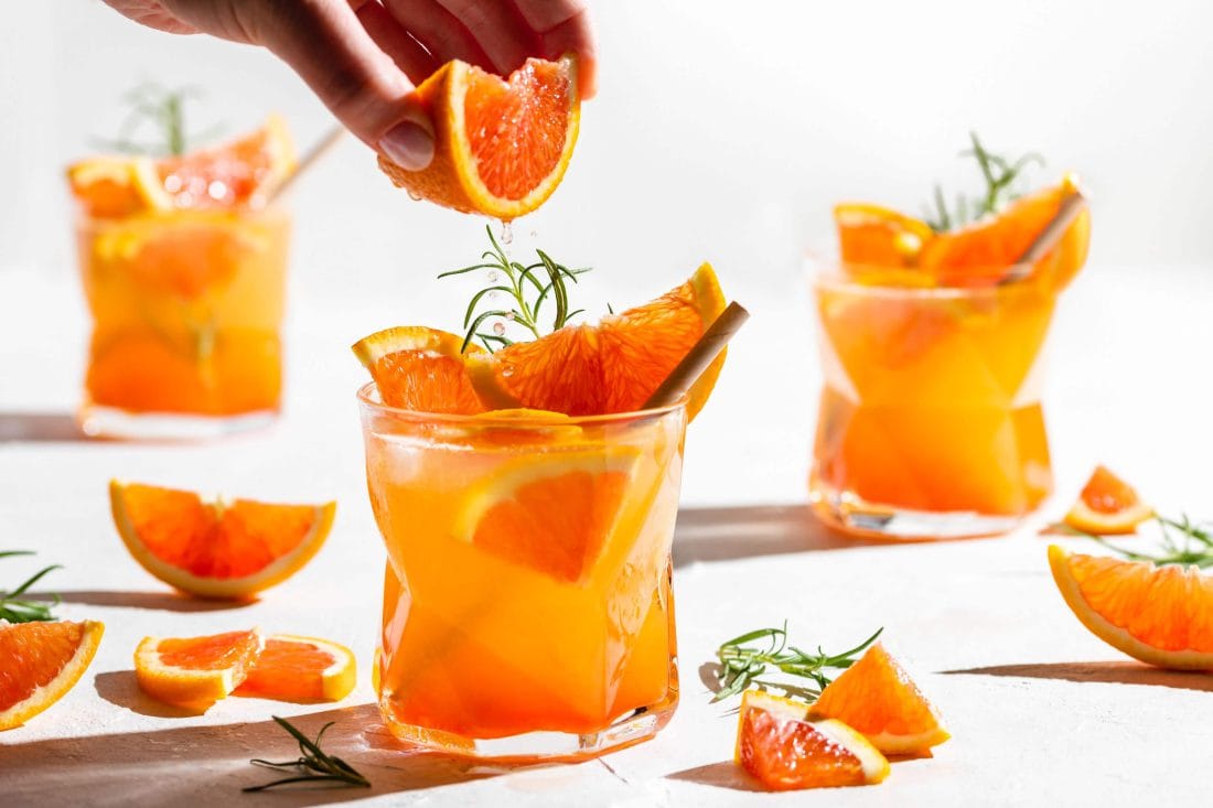 In den Sommercocktail Gin Spritz aus Aperol, Gin, Prosecco und Orangensaft wird gerade frische Orange gepresst.