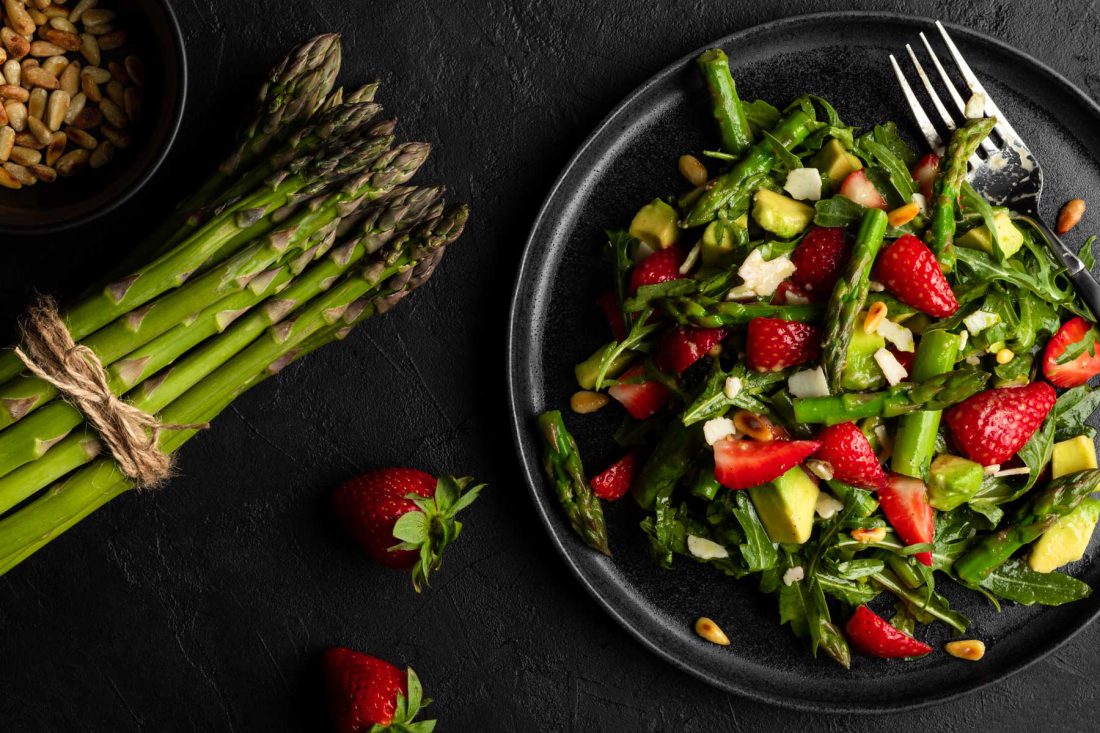 Grüner Spargel-Salat mit Erdbeeren, Rucola, Avocado, Parmesan und Pinienkernen angerichtet auf einem schwarzen Teller.