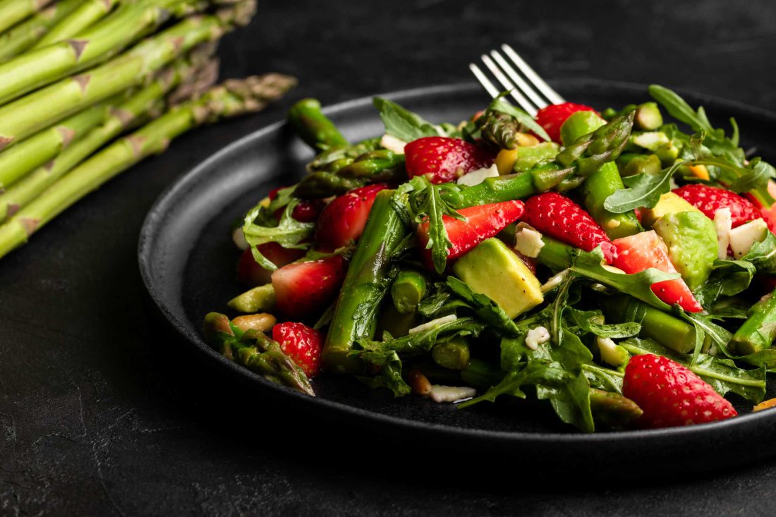 Grüner Spargel-Salat mit Erdbeeren, Avocado, frischem Rucola und Pinienkernen auf einem schwarzen Teller mit einer Gabel.