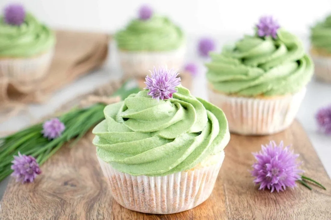 Herzhafte Muffins mit Frischkäse-Frosting auf einem Holzbrett. Neben den herzhaften Muffins liegt ein Bund Schnittlauch und Schnittlauchblüten.