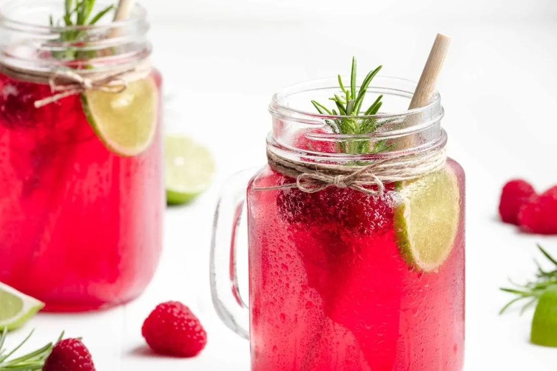 Selbstgemachte Himbeer-Limonade in zwei Gläsern, garniert mit frischem Rosmarin, Limettenscheiben und frischen Himbeeren.
