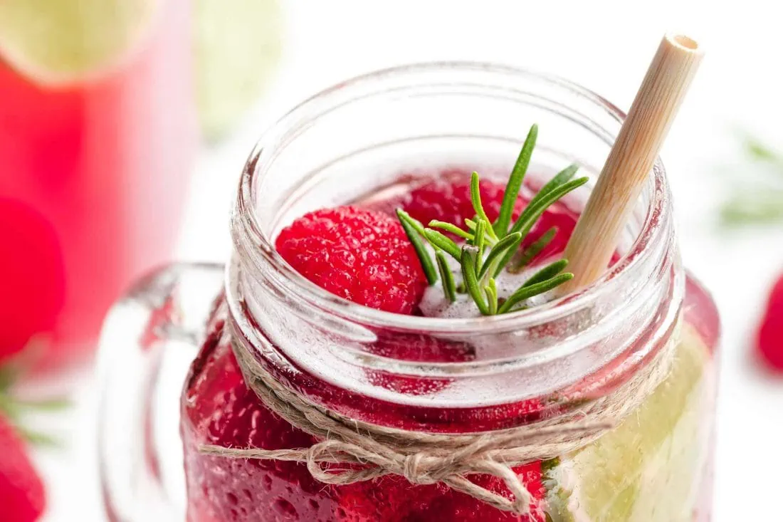 Himbeer-Limonade in einem Glas mit Strohhalm, Himbeeren, einer Limettenscheibe und einem Zweig frischem Rosmarin.