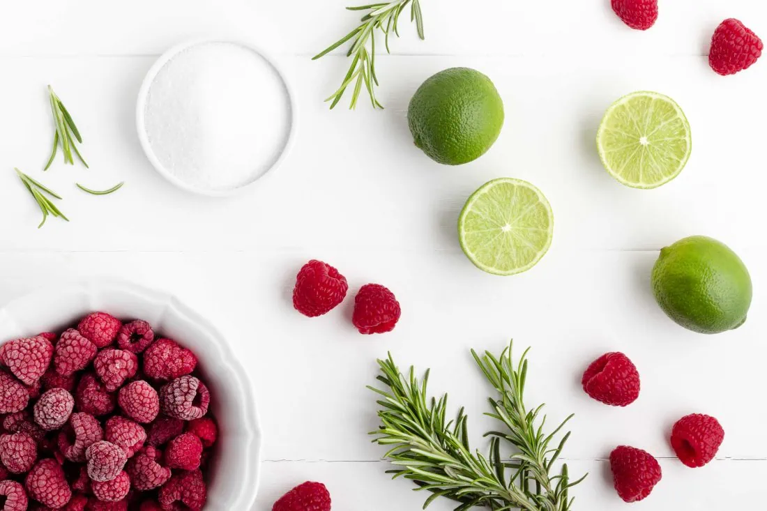 Alle Zutaten für das Rezept der selbstgemachten Himbeer-Limonade im Überblick. Darunter sind frische und gefrorene Himbeeren, Zucker, Rosmarin und Limetten.