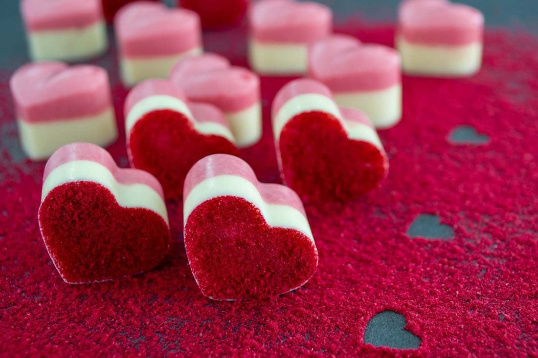 Himbeerpralinen in Herzform auf einem mit Himbeerpulver bestäubten Untergrund. Die Pralinen bestehen zur Hälfte aus weißer und rosafarbener Schokolade.