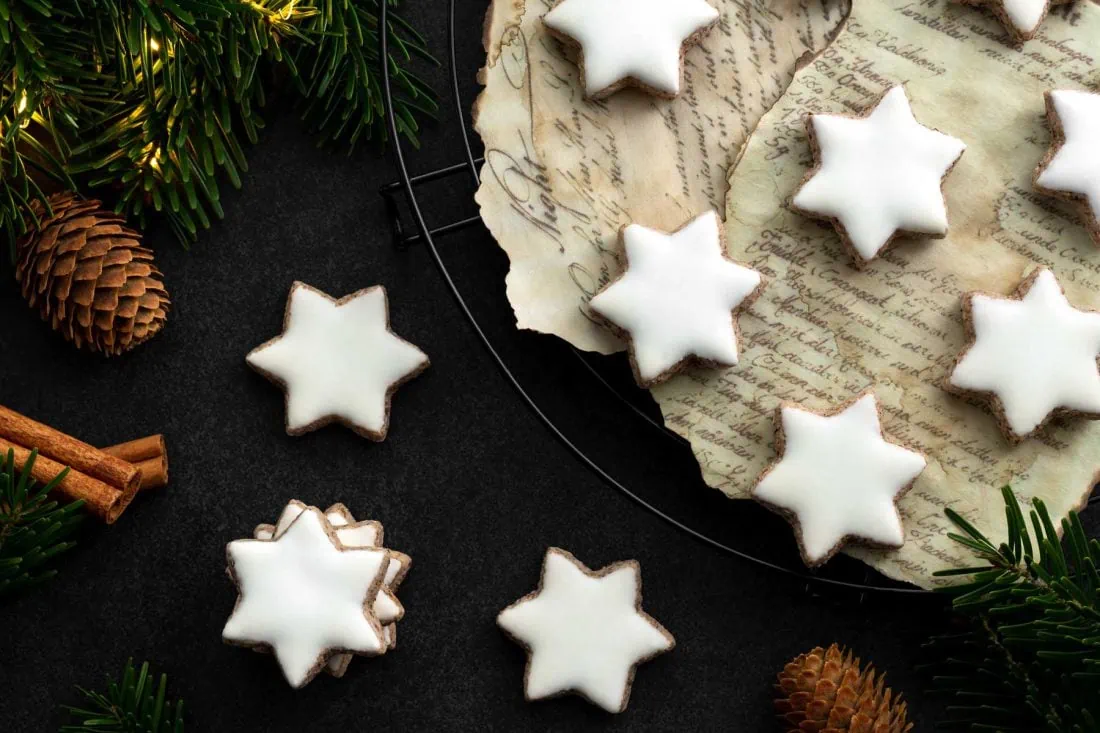 Klassische Zimtsterne auf einem Abkühlgitter. Neben den Zimtsternen liegen Tannenzweige und eine Lichterkette.
