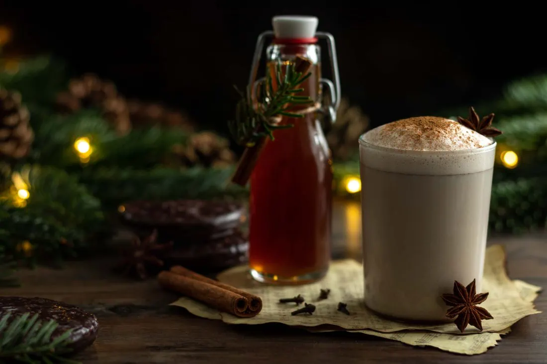 Lebkuchensirup in einer heißen, aufgeschäumten Milch, bestreut mit etwas gemahlenen Zimt. Dahinter ist eine Flasche des Sirups, Tannenzweige und eine Lichterkette.