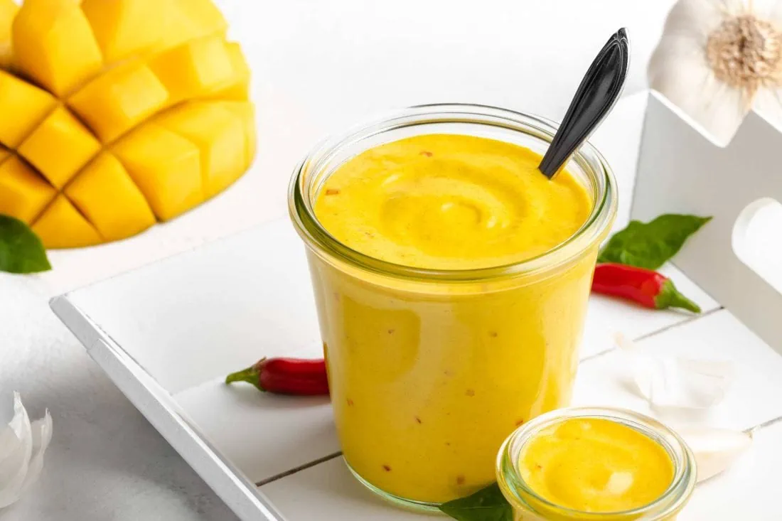 Mango Chili Aioli in einem Glas mit einer aufgefächerten Mango und roten Chilischoten dahinter.