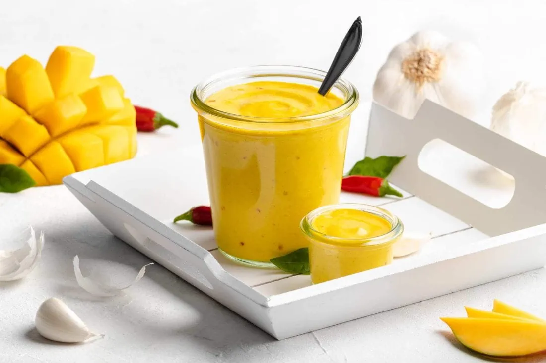 Mango Chili Aioli in zwei Gläsern auf einem weißen Tablett. Daneben liegen Chilis, Knoblauch und Mangos.