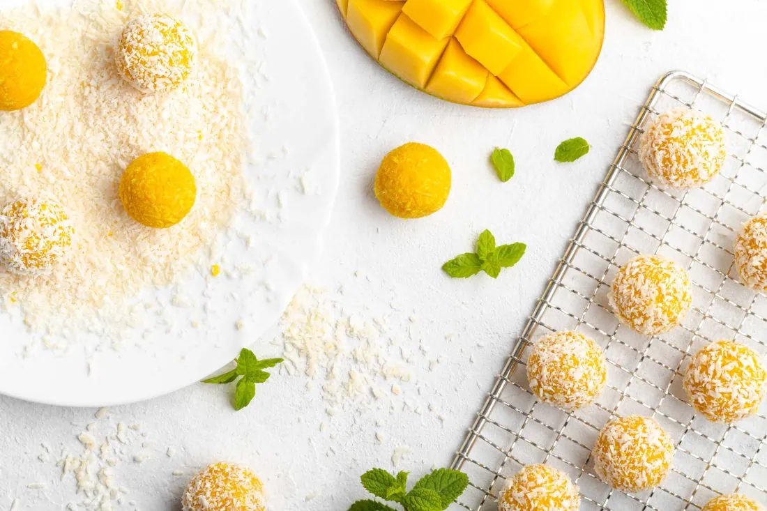 Mango Ladoo werden auf einem Teller in Kokosraspeln gewälzt. Neben der indischen Süßigkeit liegen eine Mango und Minze.