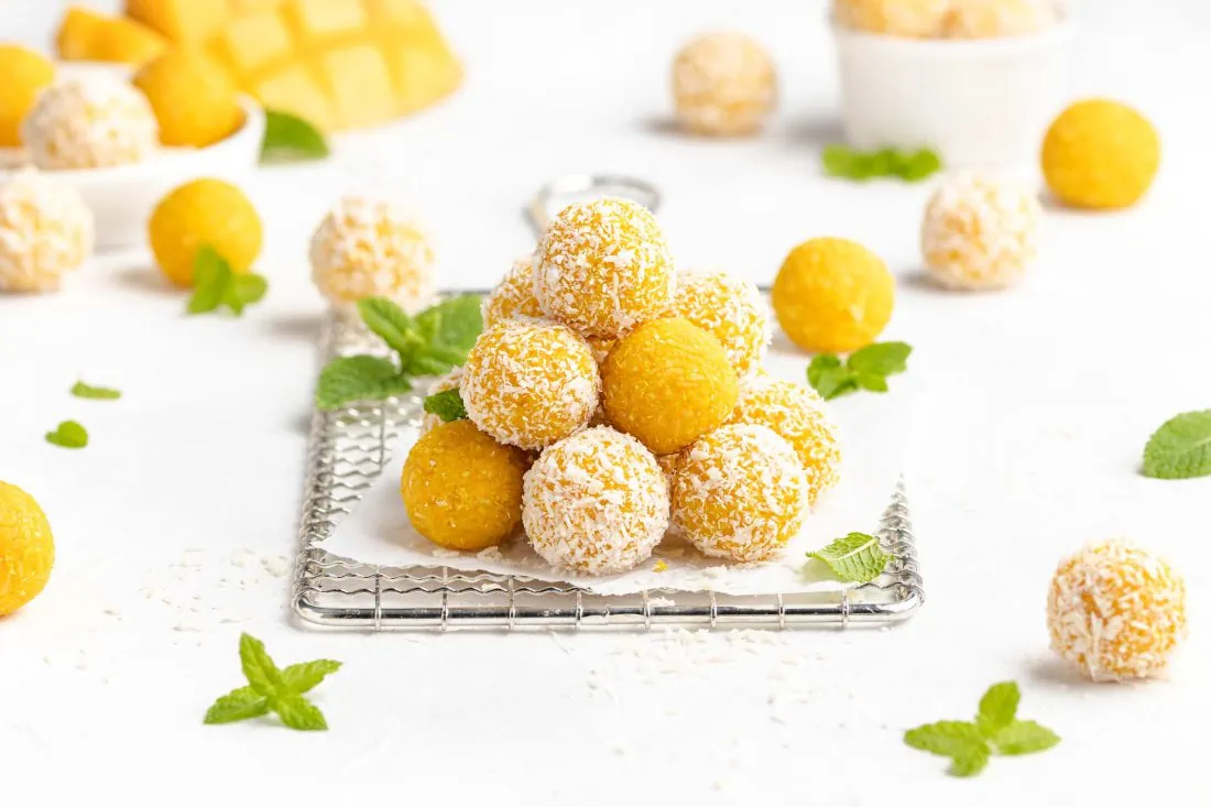 Mango Ladoo zu einem Türmchen gestapelt auf einem weißen Papier. Neben der indischen Süßigkeit liegen Kokosraspeln und frische Minze.