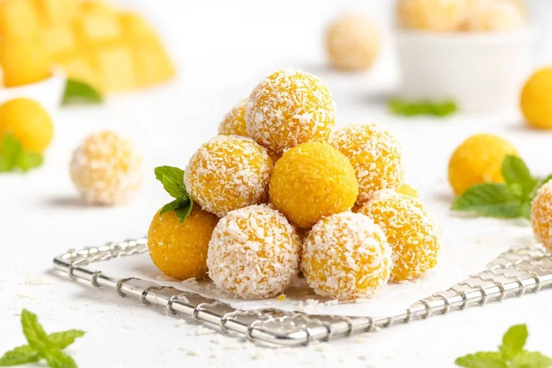 Mehrere Mango Ladoo, zum Teil umhüllt mit Kokosraspeln, aufeinander gestapelt auf einem Gitter. Daneben liegen frische Minze und Mango.