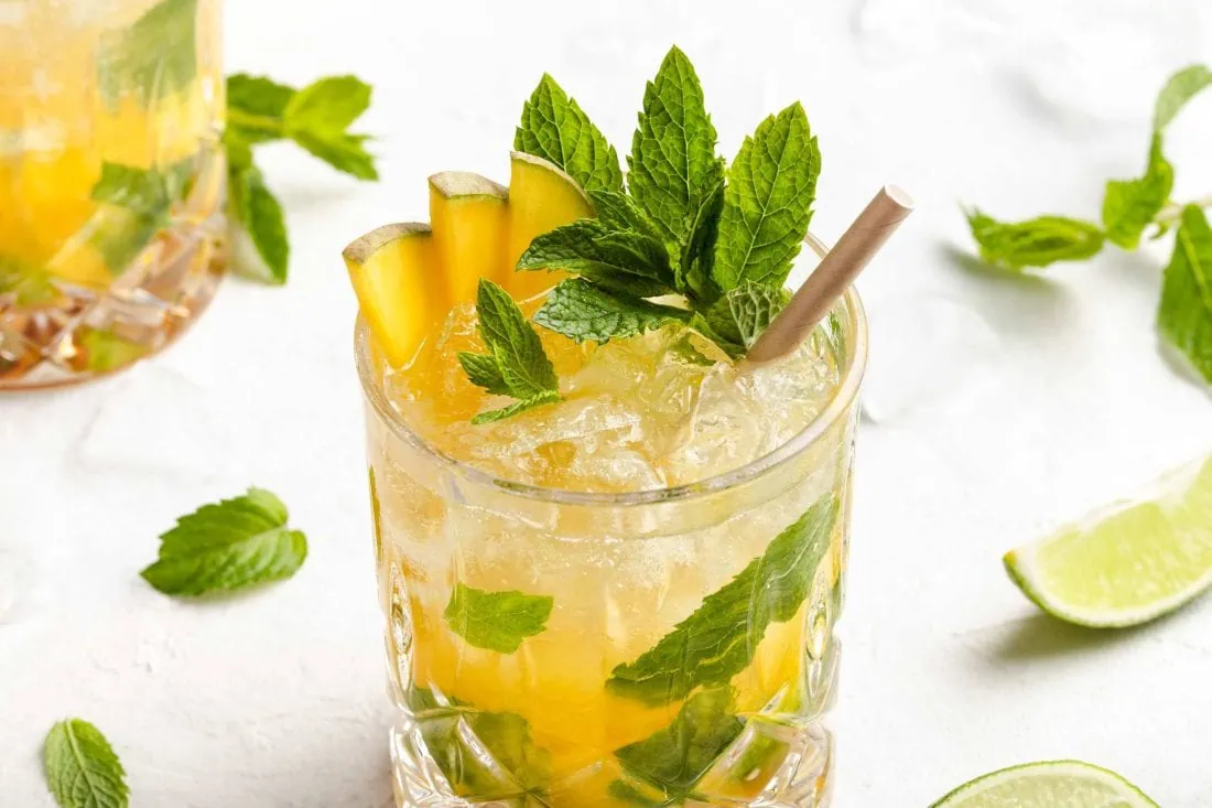 Mango Mojito in einem Tumbler mit Strohhalm, einem Zweig frischer Minze und aufgeschnittener Mango.