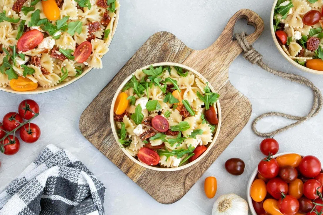 Mediterraner Nudelsalat verteilt auf drei Schüsseln. Um den Nudelsalat herum liegen mehrere Tomaten, ein Geschirrhandtuch und eine Knoblauchzehe.