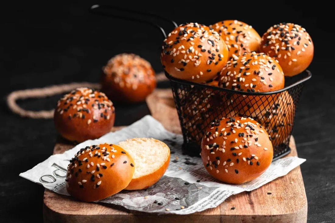 Mini Brioche Burger Buns mit schwarzem und weißem Sesam in einem kleinen Körbchen und aufgeschnitten auf einem Holzbrett.