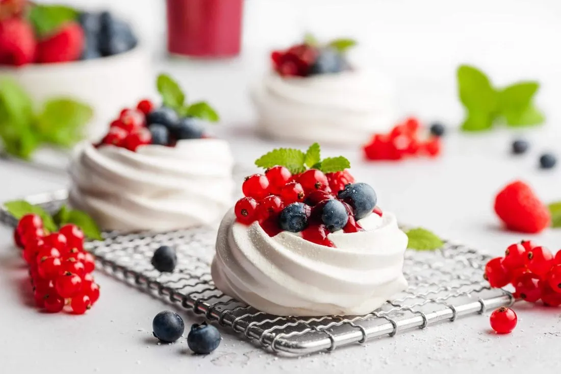 Mini Pavlova Dessert mit Mascarpone und frischen Beeren. Neben den Pavlova liegen Blaubeeren und Johannisbeeren.