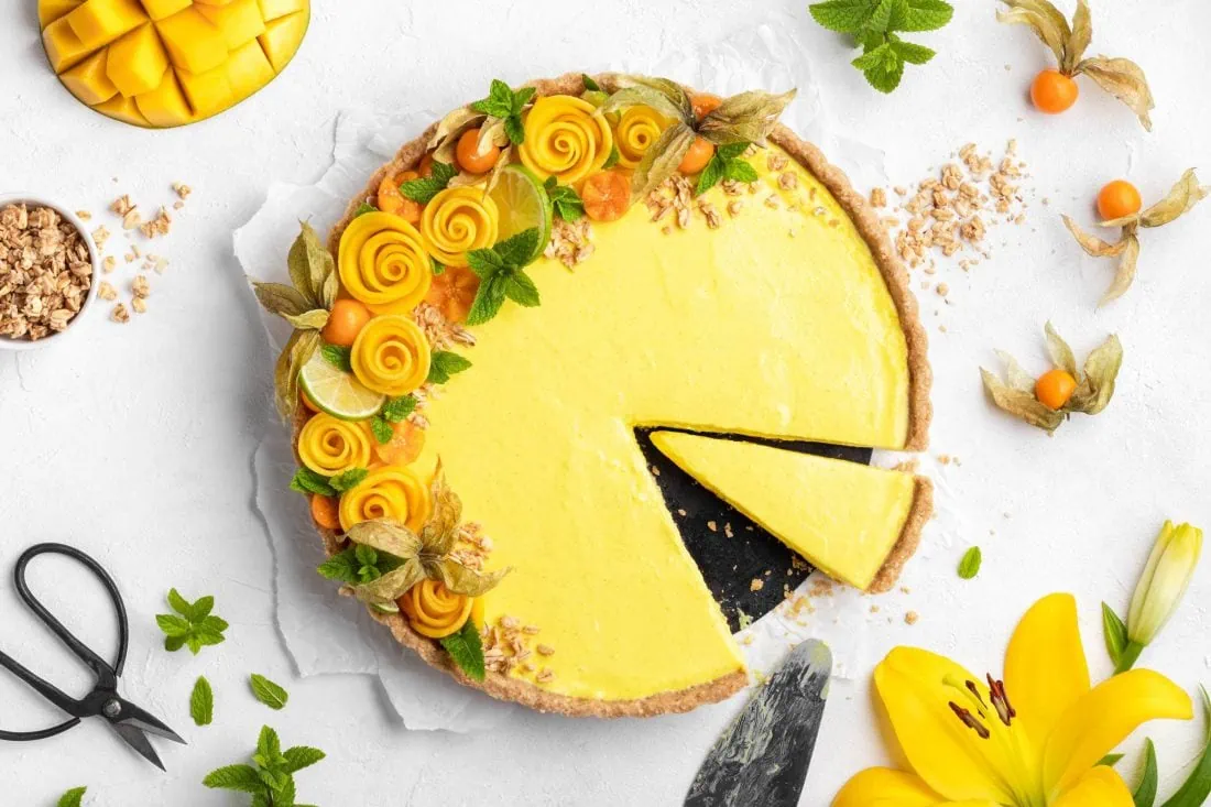 No Bake Tarte mit Mango angeschnitten auf einem weißen Untergrund sommerlich garniert. Neben der Tarte liegen mehrere Zutaten.