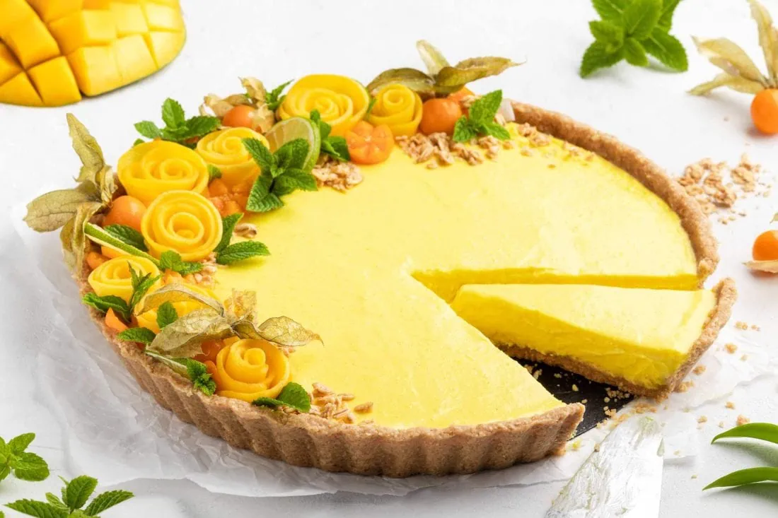 No Bake Mango Tarte angeschnitten auf einem Teller. Hinter der Tarte liegen Physalis und eine aufgefächerte Mango.