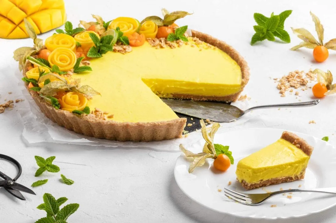 Ein Stück sommerliche No Bake Mango Tarte auf einem Teller mit einer Gabel. Im Hintergrund steht die restliche angeschnittene Tarte.