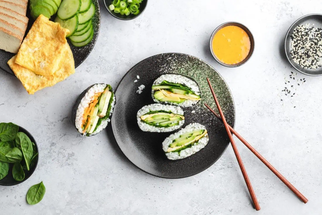 Mehrere Onigirazu auf einem Teller, daneben liegen die geschnittenen Zutaten für die Füllung der Sushi Sandwichs.