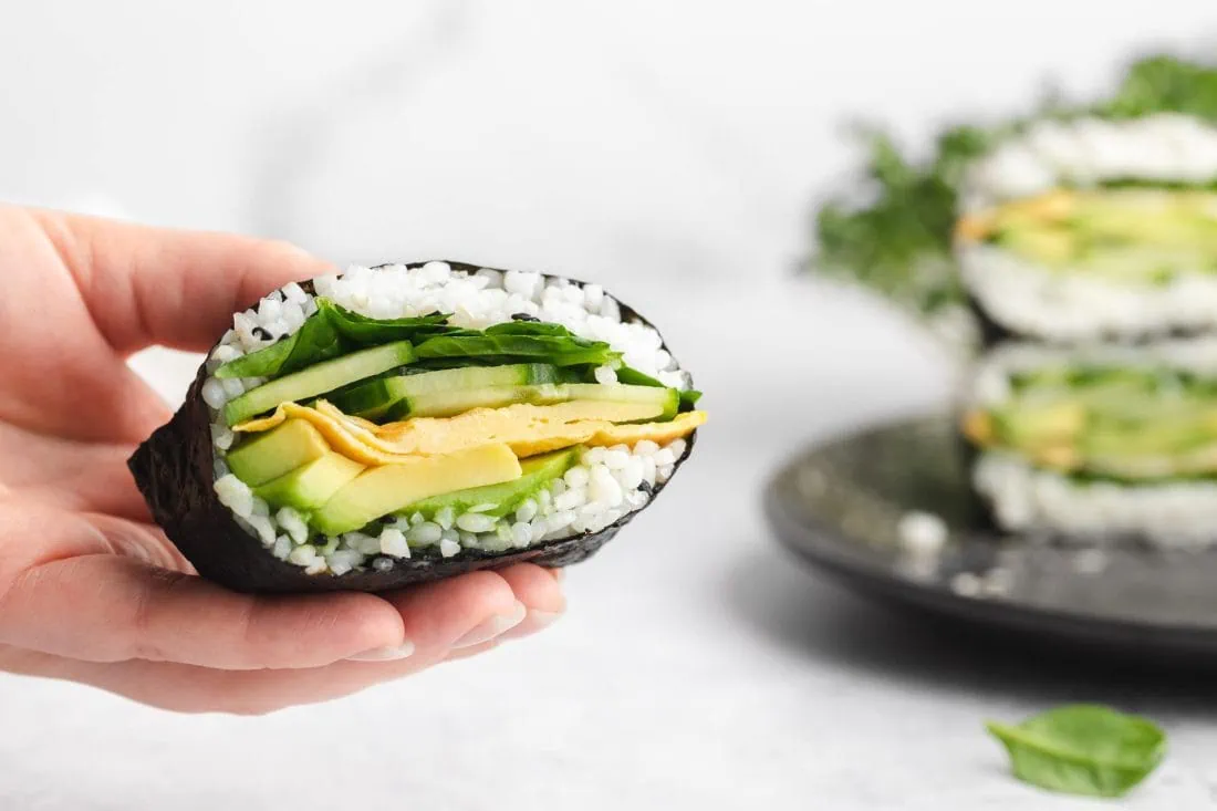 Ein Onigirazu wird in einer Hand gehalten. Dahinter liegen weitere halbierte Sushi Sandwichs.