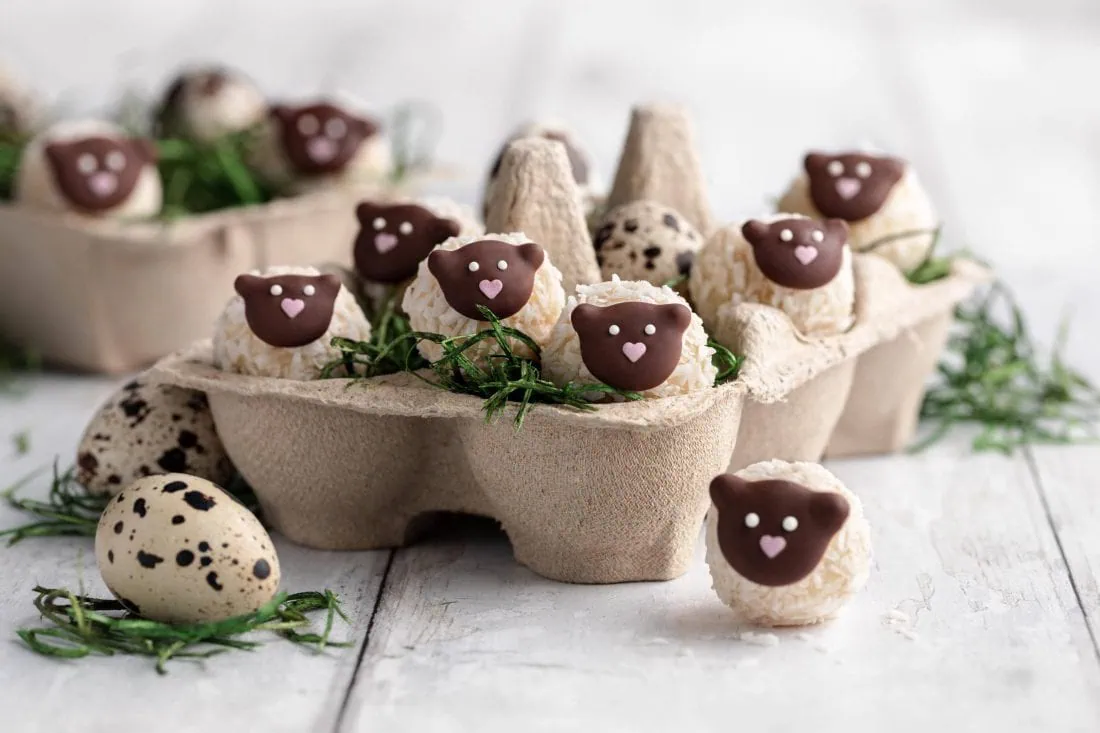 Mehrere Schäfchen Pralinen in einem einem Eierkarton als Osternest. Die Osterpralinen bestehen aus weißer Schokolade und Kokosraspeln.