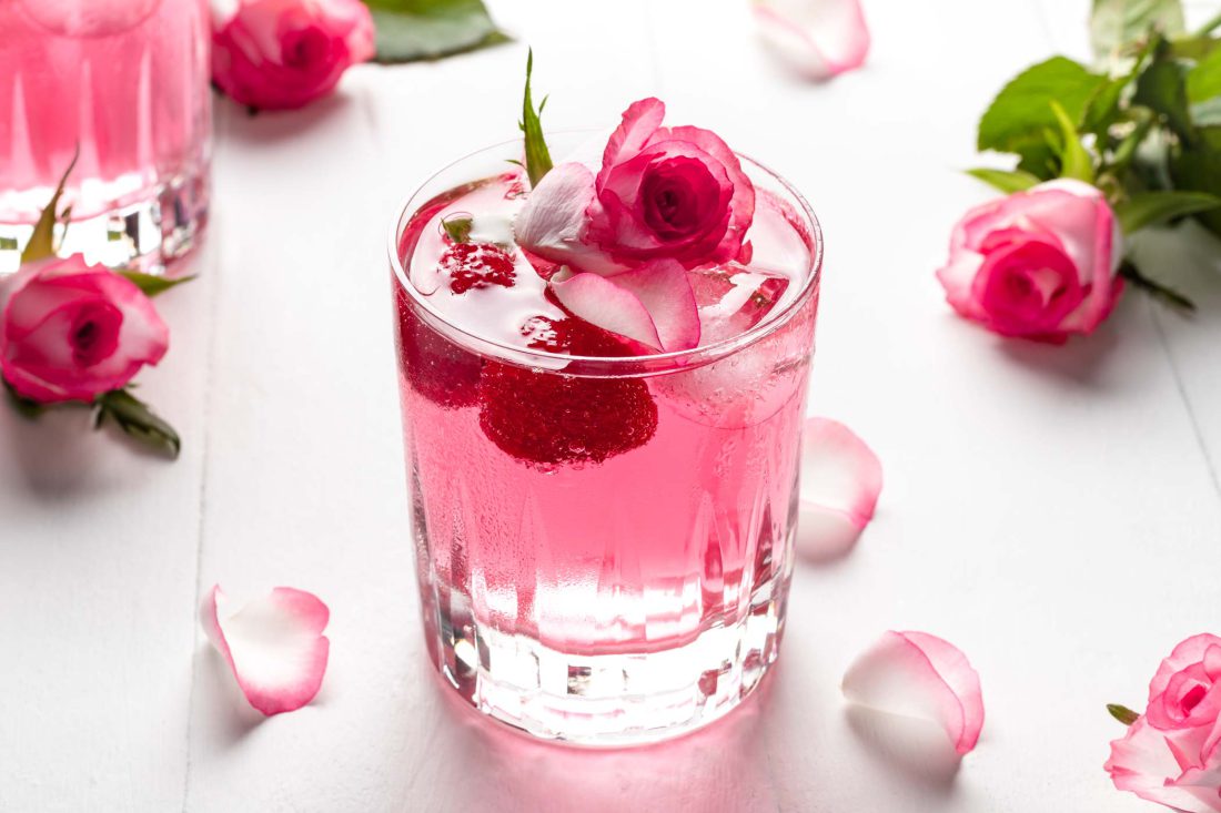 Pinker Gin Wild Berry mit Rosenwasser und Himbeeren auf einem hellen Untergrund umgeben von Rosenblütenblättern.