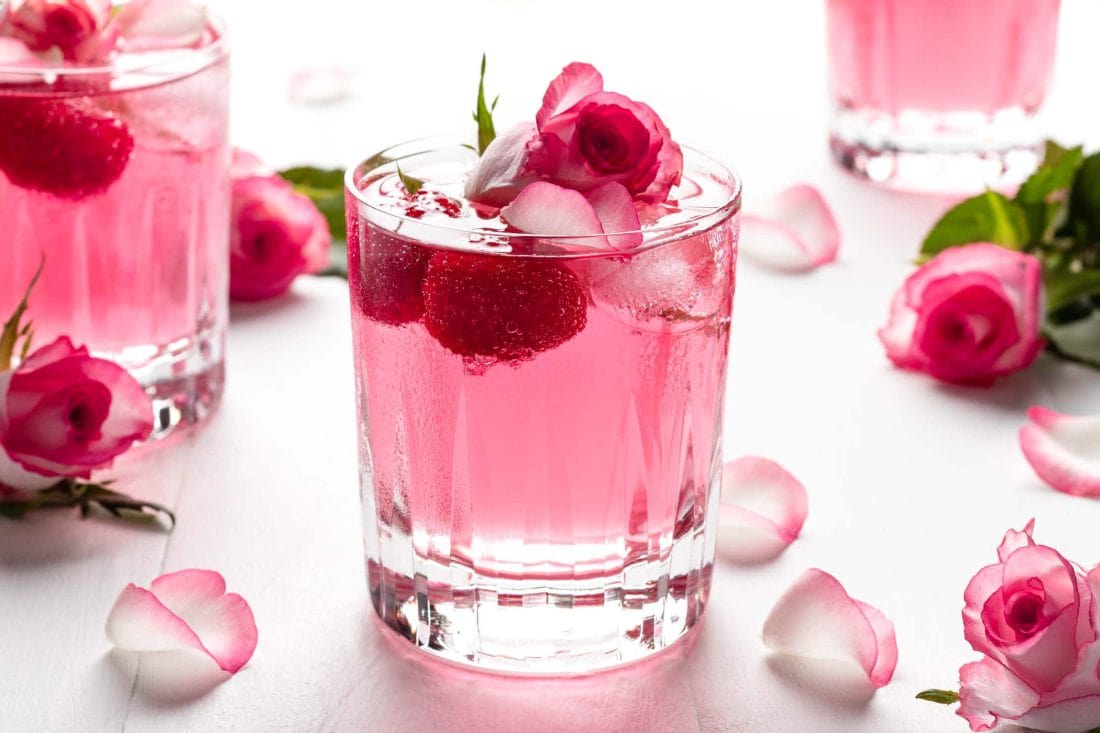 Pinker Gin Wild Berry mit Rosenwasser und Himbeeren in einem kleinen Glas. Neben dem Glas liegen Rosen und Rosenblütenblätter.