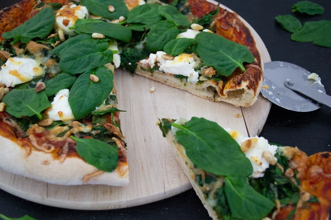 Eine knusprig gebackene Pizza belegt mit Ricotta und frischem Spinat, überbacken mit Käse, liegt auf einem hellen Holzbrett. Ein abgeschnittenes Stück wird gerade vom Brett genommen.