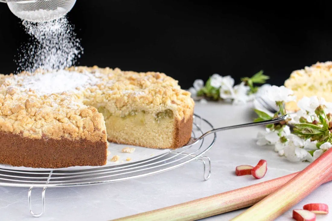 Auf einem runden Abkühlgitter liegt ein angeschnittener Rhabarberkuchen mit Streuseln. Der Rhabarberkuchen wird gerade mit einem feinen Sieb mit Puderzucker bestreut.