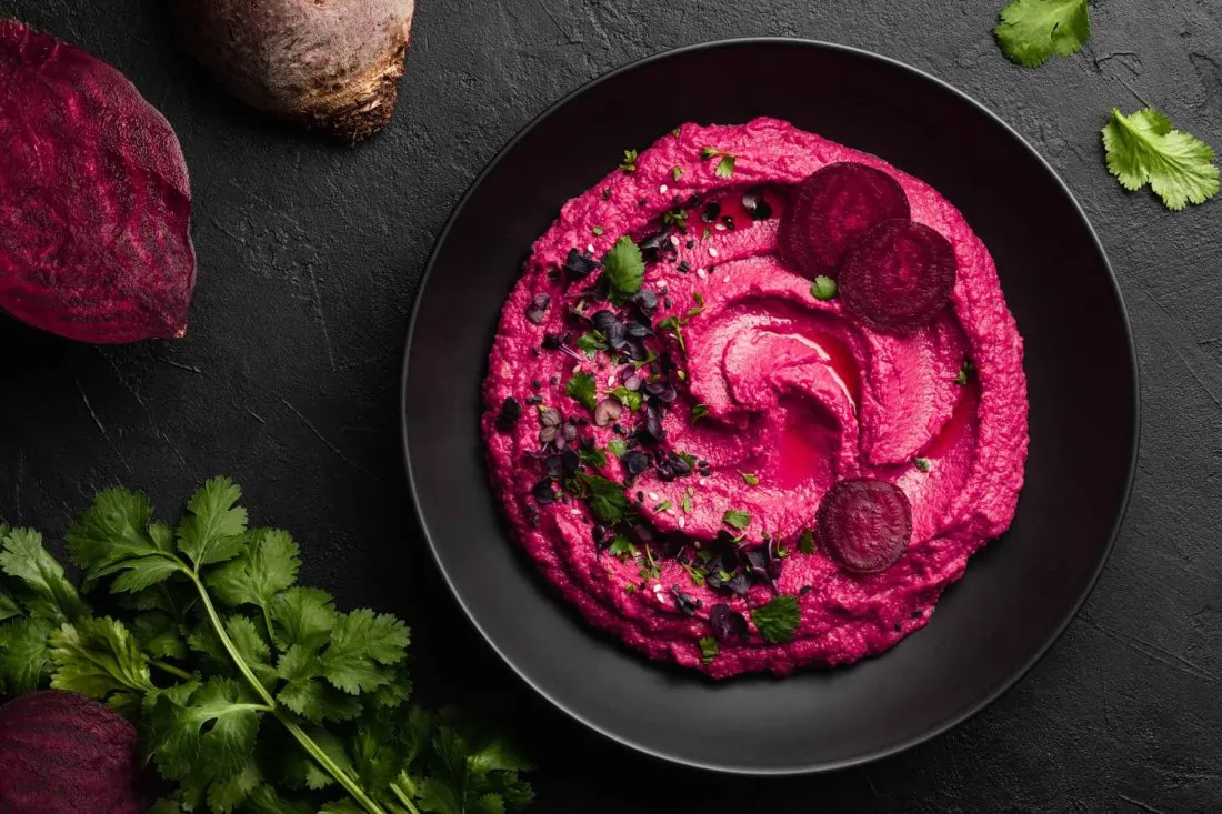 Rote Bete Hummus in einem schwarzen Teller. Neben dem Teller liegt frische, aufgeschnittene Rote Bete.