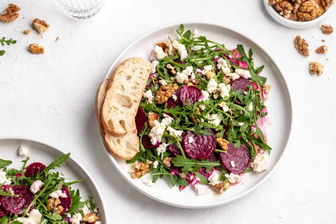 Rote Bete Salat mit Schafskäse und Walnüssen angerichtet auf einem weißen Teller und serviert mit zwei Scheiben Brot.