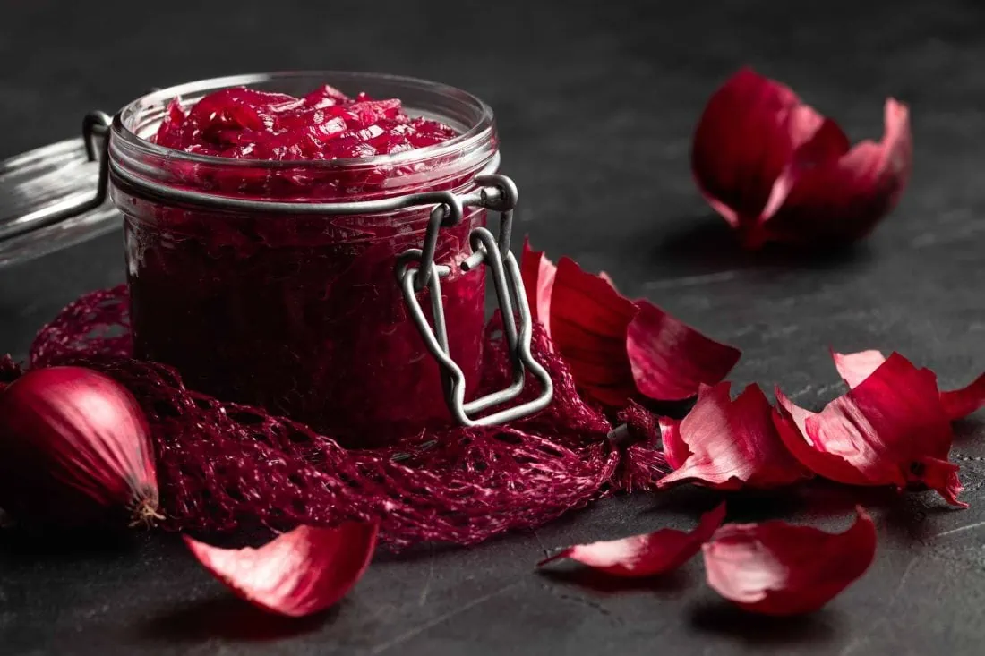Rotes Zwiebel Chutney in einem Bügelverschlussglas umgeben von den Schalen mehrerer roter Zwiebeln.
