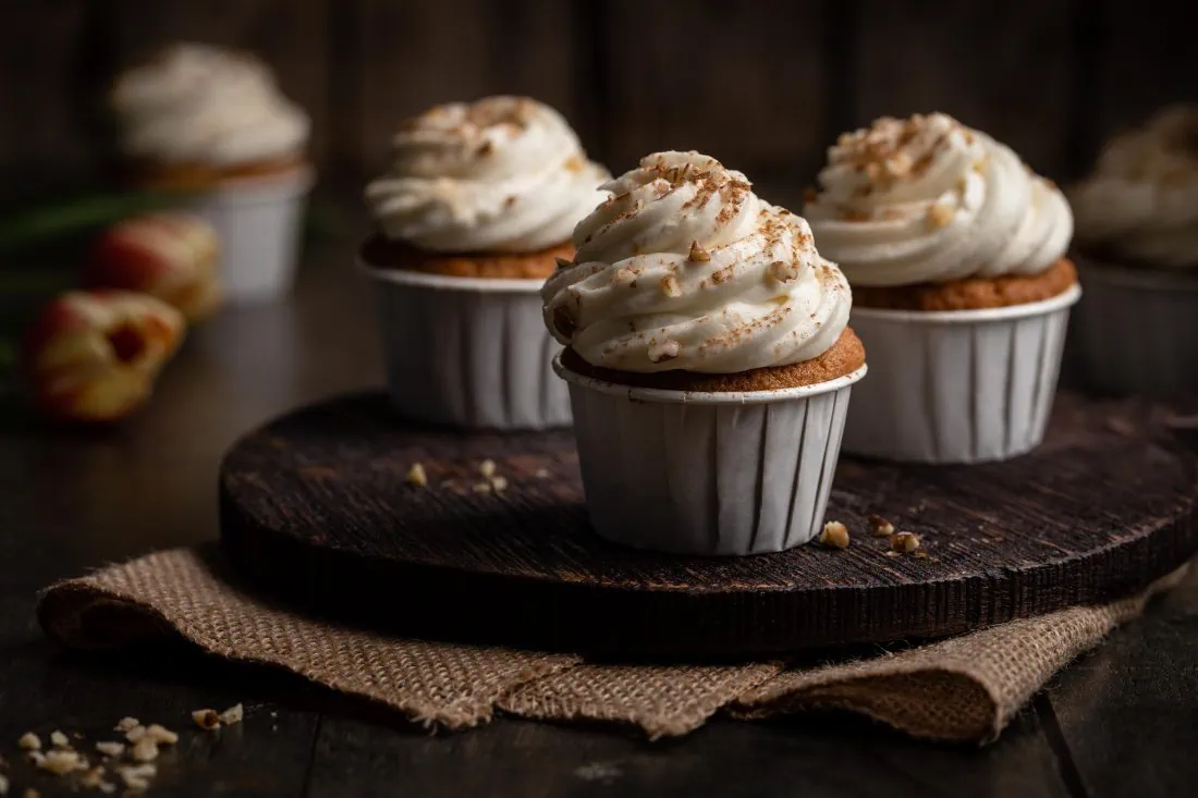 Mehrere saftige Karotten Cupcakes in weißen Papierförmchen mit einem Frischkäse-Frosting als Topping und gehackten Walnüssen als Garnitur.