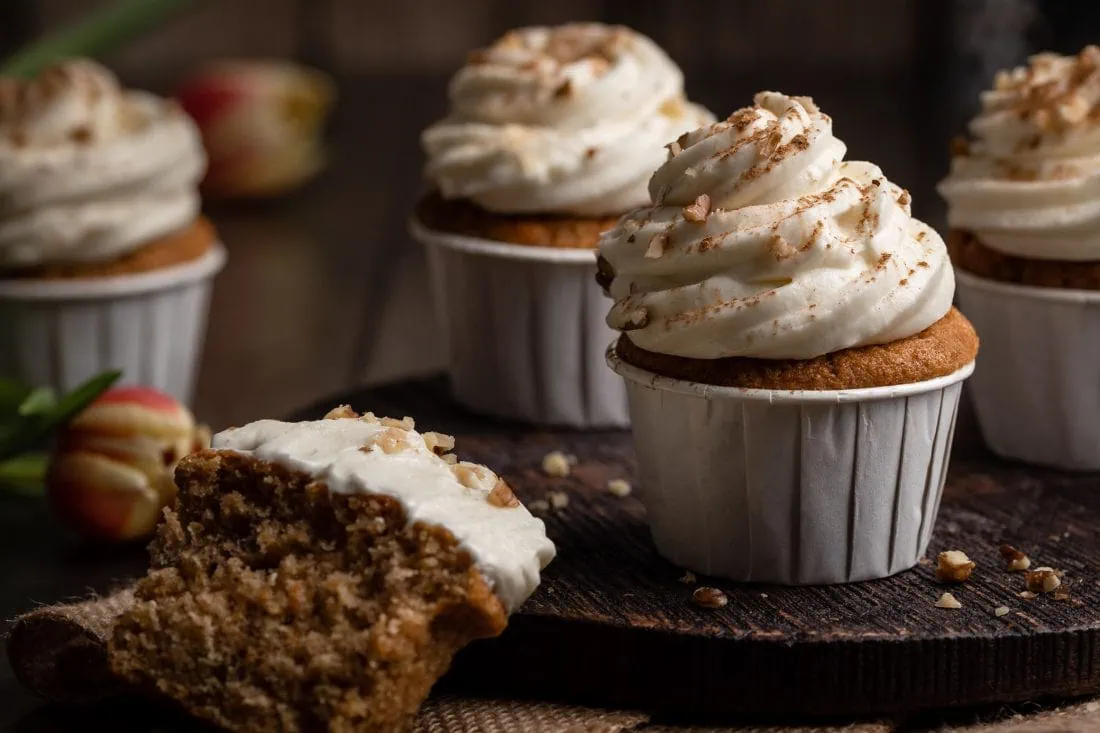Mehrere saftige Karotten Cupcakes mit Frischkäse-Frosting. Einer der Cupcakes liegt halbiert an der Seite.