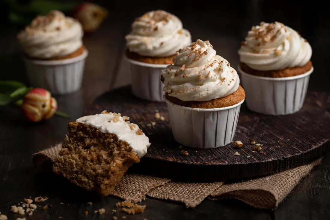 Vier Karotten Cupcakes mit Walnüssen und einem weißen Frischkäse-Frosting auf einem Holzbrett.