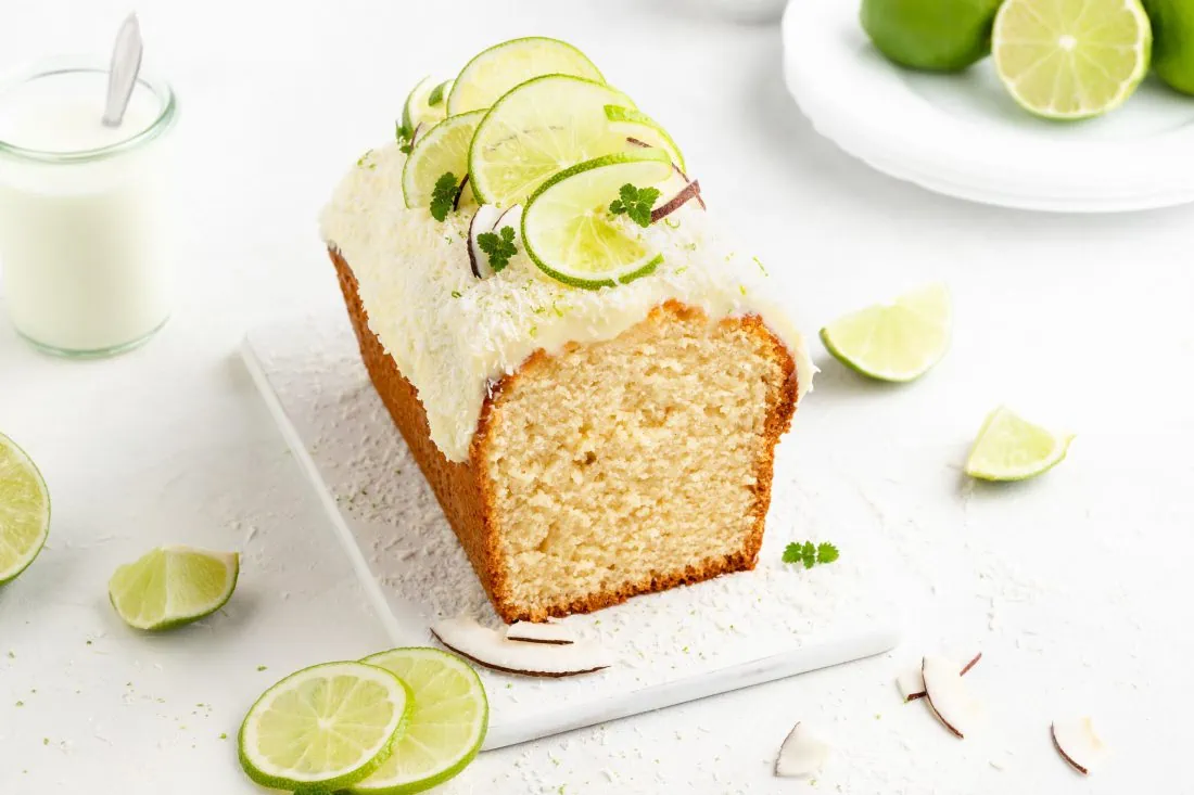 Ein Limettenkuchen mit Kokos gebacken in einer Kastenform und verfeinert mit einem weißen Frosting auf einem weißen Brett.