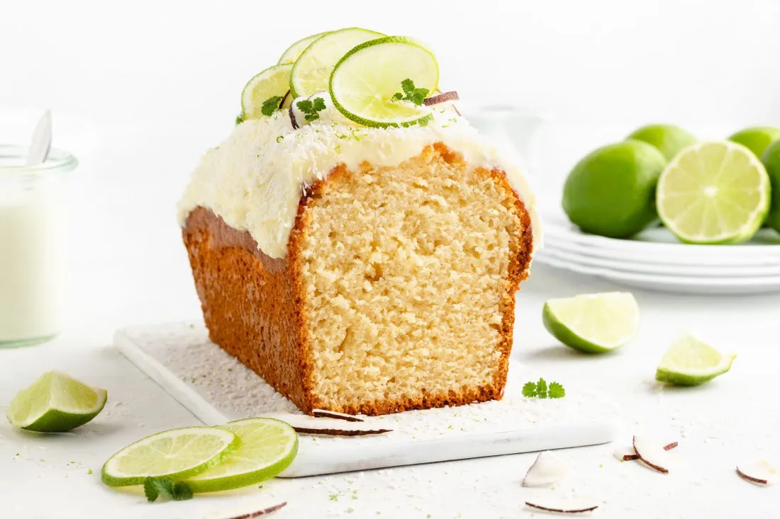 Ein angeschnittener Kokos-Limetten-Kuchen getoppt mit einem Frischkäse-Frosting und garniert mit Limettenscheiben.