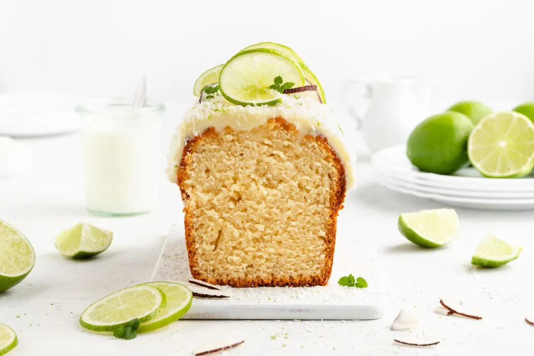 Ein angeschnittener Limettenkuchen mit Kokos auf einer weißen Platte. Daneben liegen Limtettenscheiben und Kokosraspeln.
