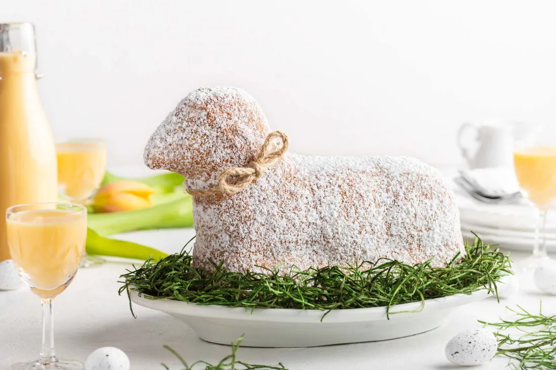 Mehrere Likörgläser mit Eierlikör und ein saftiges Osterlamm mit Puderzucker stehen auf einem weißen Untergrund.